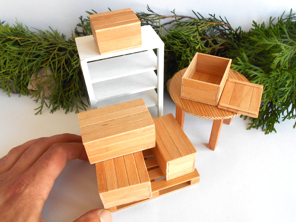 A miniature wooden toy furniture set including a white three-tier cabinet, a round table with a wooden crate, and a hand holding a matching crate and pallet, styled with green pine branches on a white background.