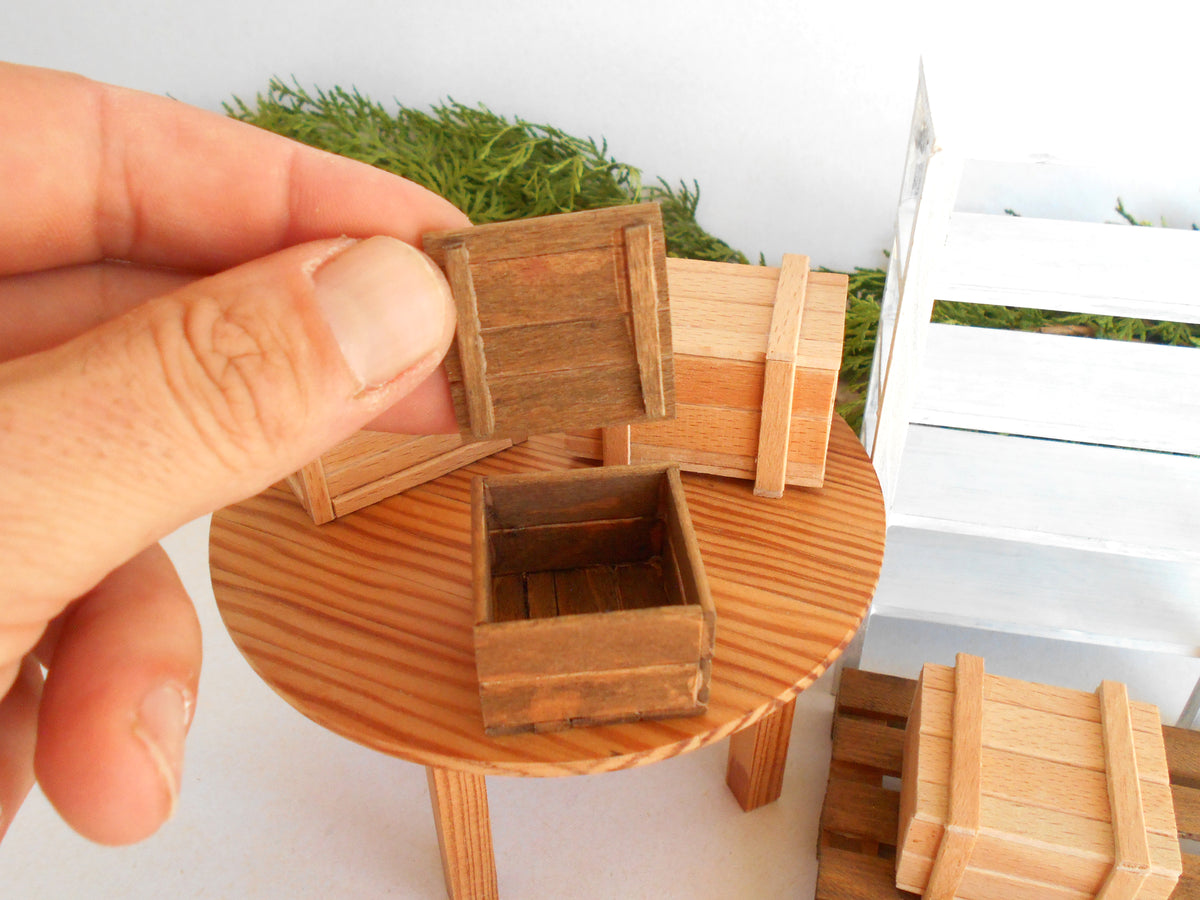 A hand holding a miniature wooden crate box, with several small wooden storage crates and a round wooden table in a dollhouse-scale setting.