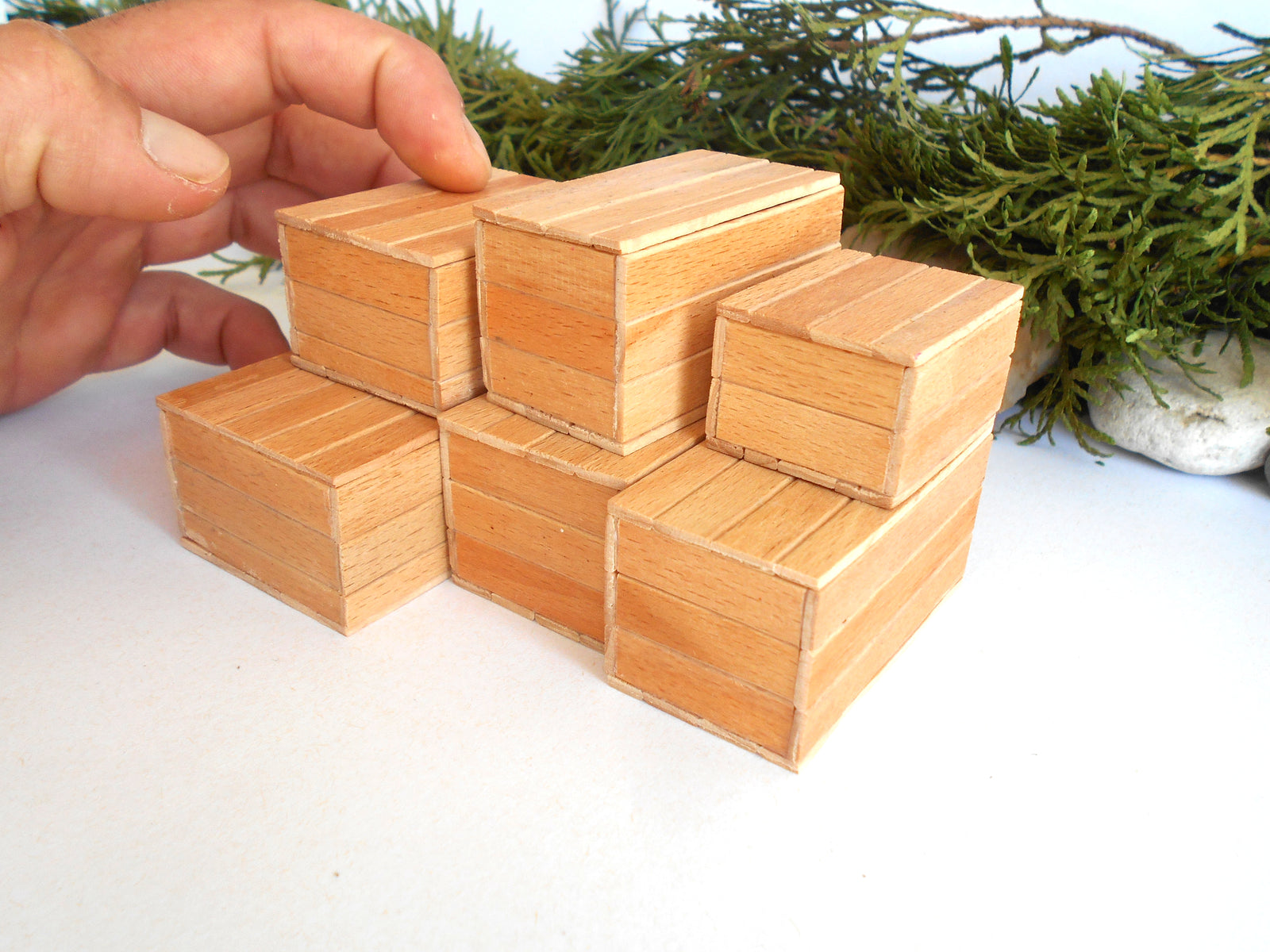 A hand is holding a small wooden miniature boxes structure made of multiple miniature light brown wood blocks, with green foliage in the background.