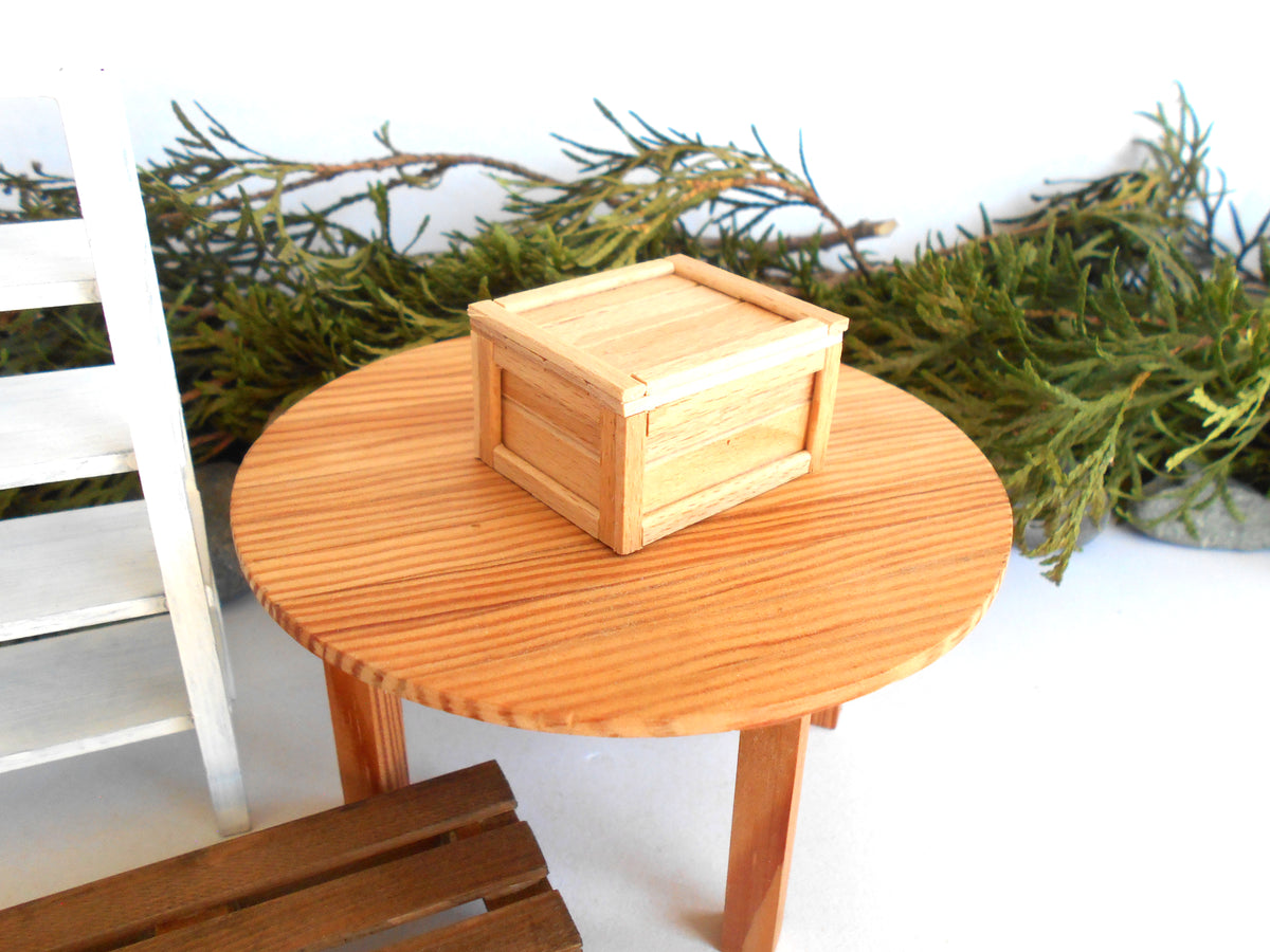 A small square wooden box with a lid sits on a round wooden table, with green branches in the background and part of a white chair visible on the left.