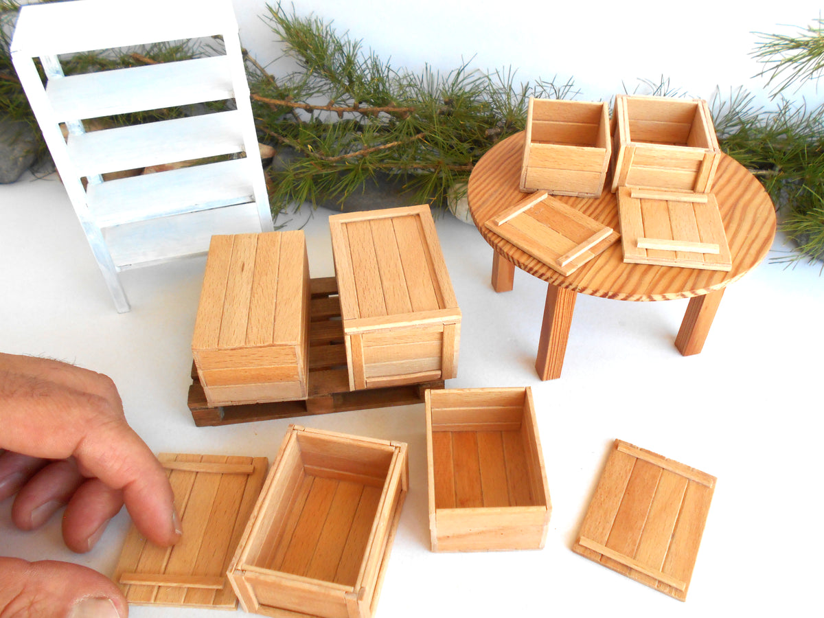 A set of miniature light wood crates and boxes for dollhouse or model building, shown with a hand adjusting one piece and a small round table with matching pieces in the background.