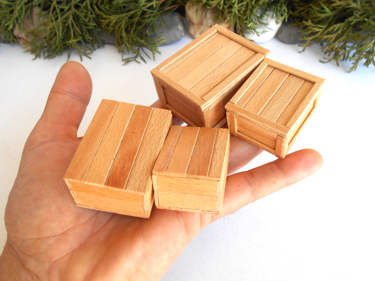 Four miniature light brown wooden boxes with a plank design, held in a person's hand to show scale against a white background with green foliage at the top.