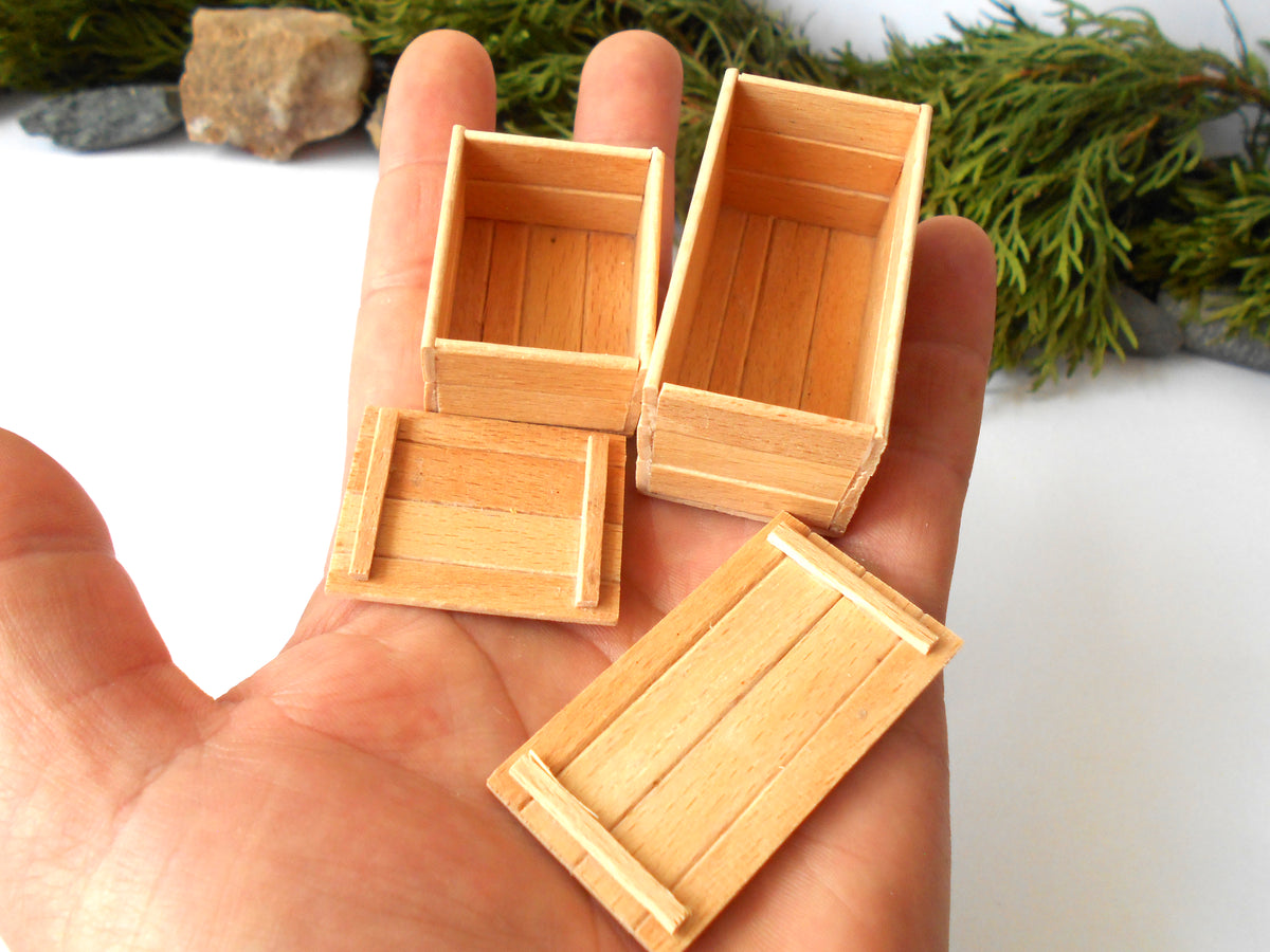 Four miniature light wood boxes of different sizes held in a person's hand, with a white background and some greenery and rocks at the top.