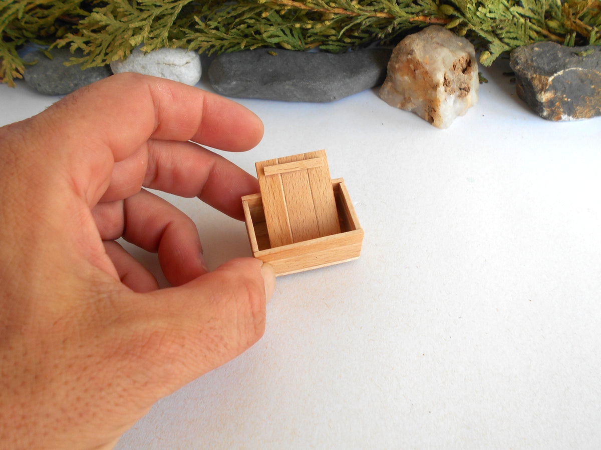 A miniature light wood box or miniature furniture piece being held by a person's hand against a white background with decorative rocks and greenery.