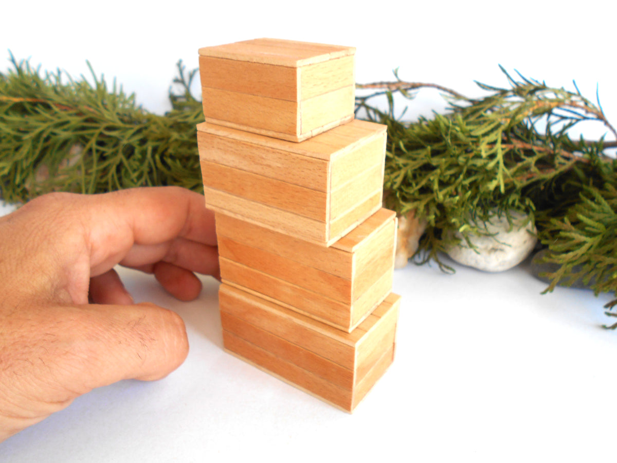 A hand is adjusting a stack of four mini, light-colored wooden boxes with a natural wood grain finish, set against a white background with green pine branches in the background.