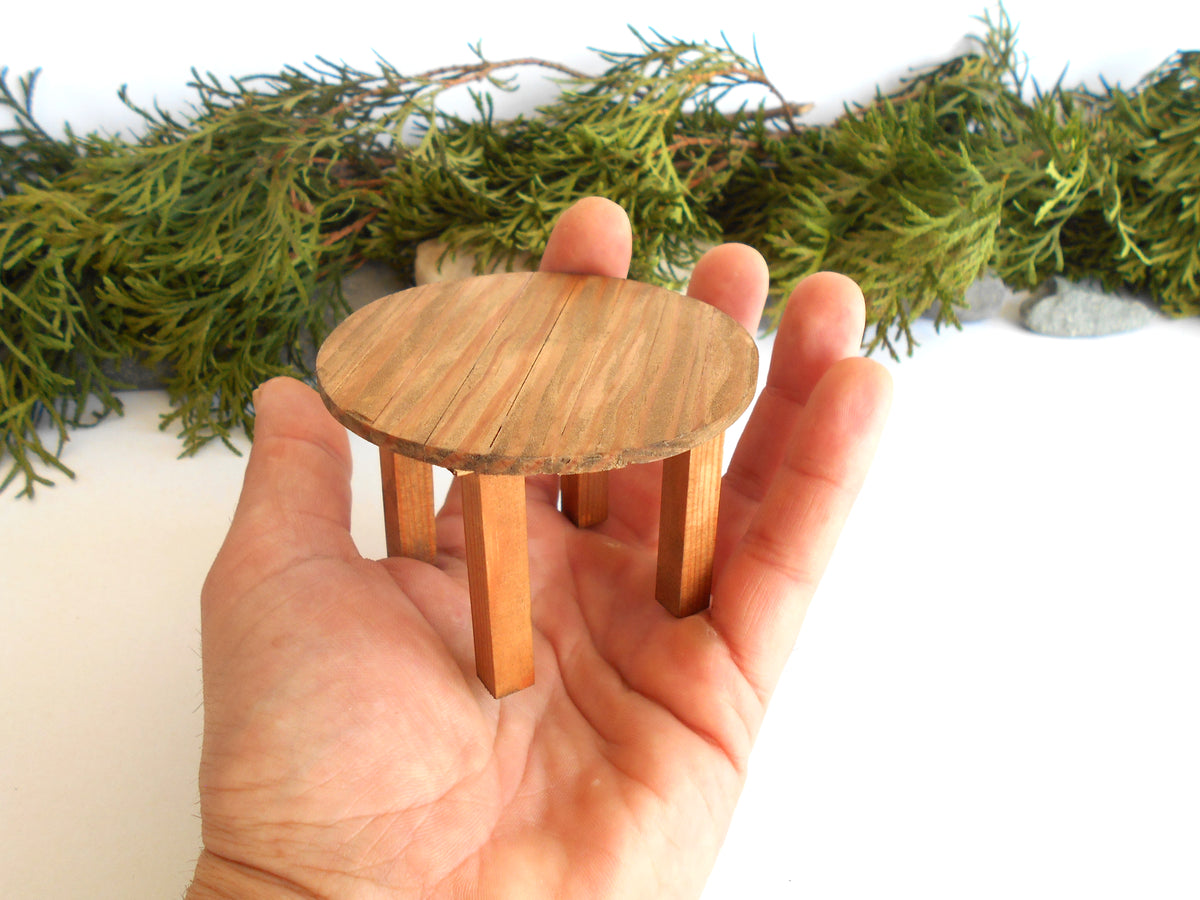 A miniature round wooden stool or table with four legs, held in a person's hand to show its small scale, against a white background with green foliage garland.