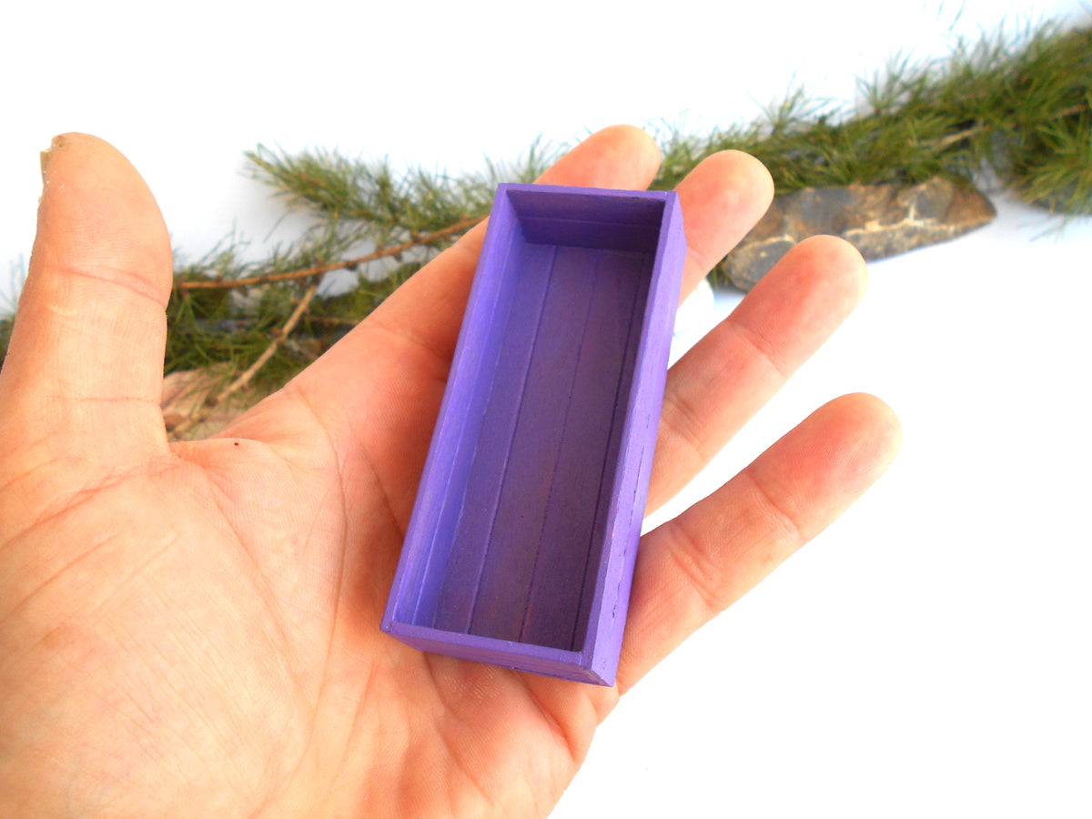 A hand holding a small rectangular purple box with a ribbed interior, shown against a white background with some green pine branches in the upper area.