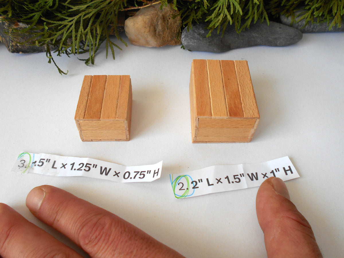Two small wooden boxes are shown on a white surface with printed size strips below them and a person's fingers pointing at the text. Greenery and stones decorate the background.