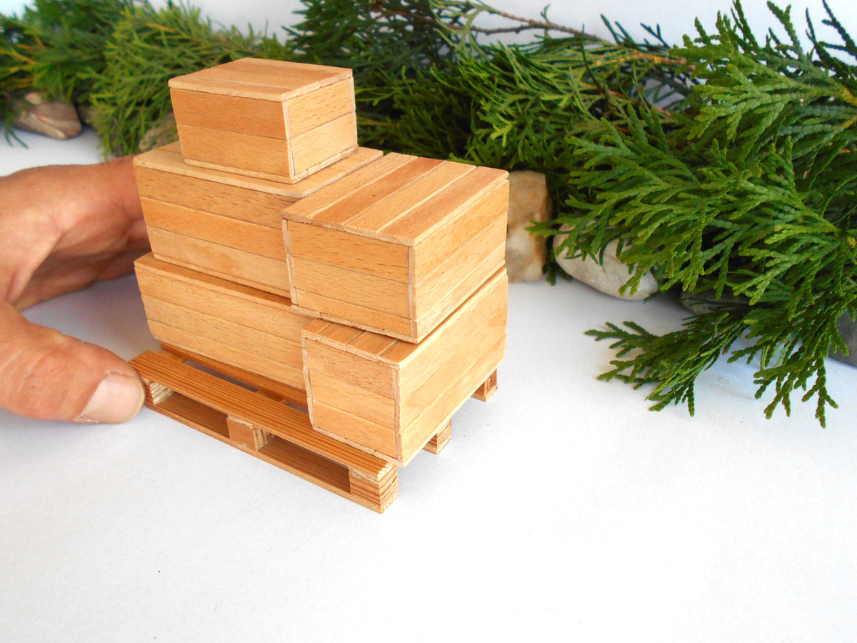 A miniature wooden toy stack of stacked boxes on a small wooden pallet, held by a person's hand against a white background with green foliage.