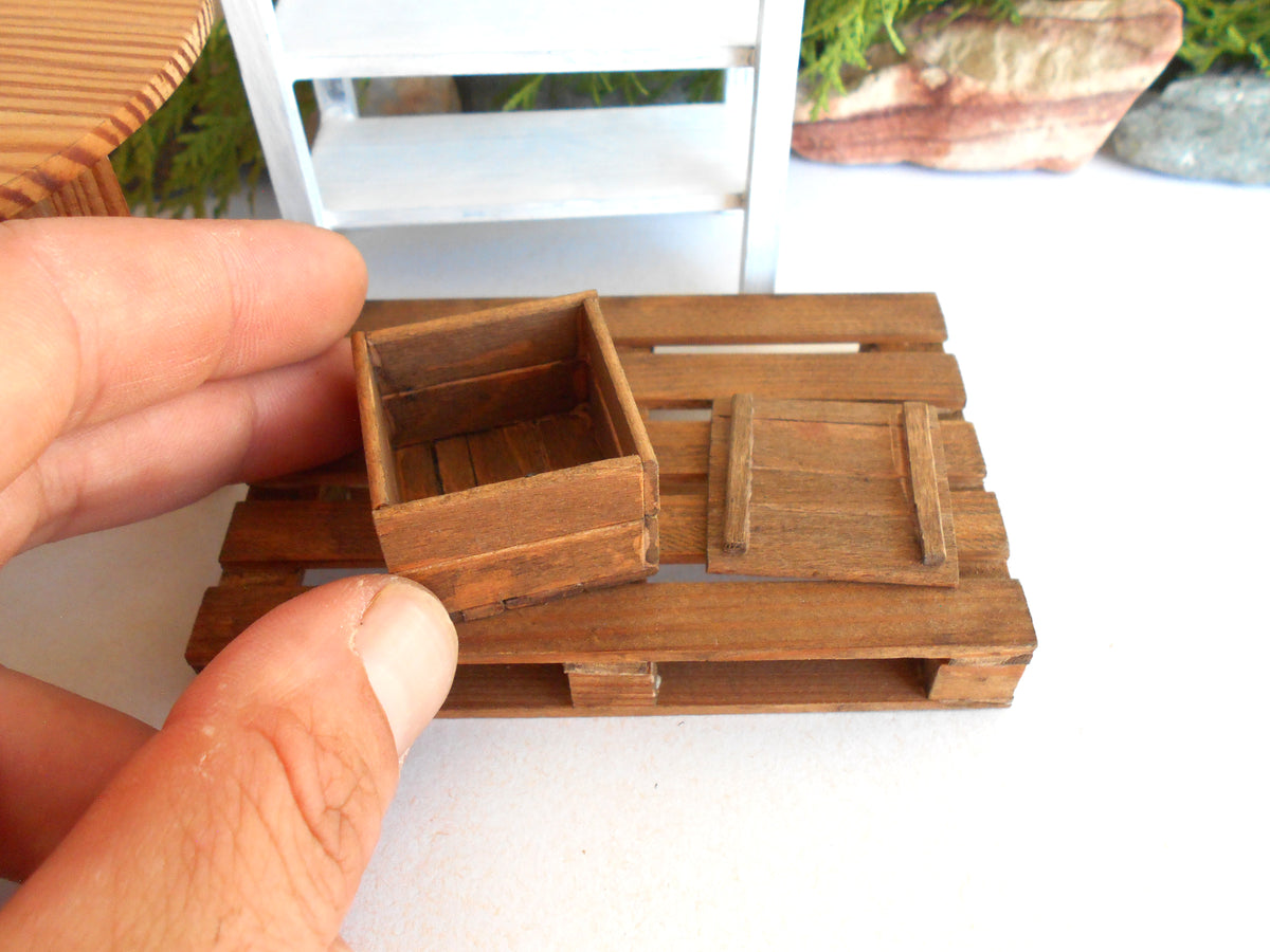 A miniature wooden crate and matching lid set held by a person's hand, shown in close-up on a white surface with a blurred background.