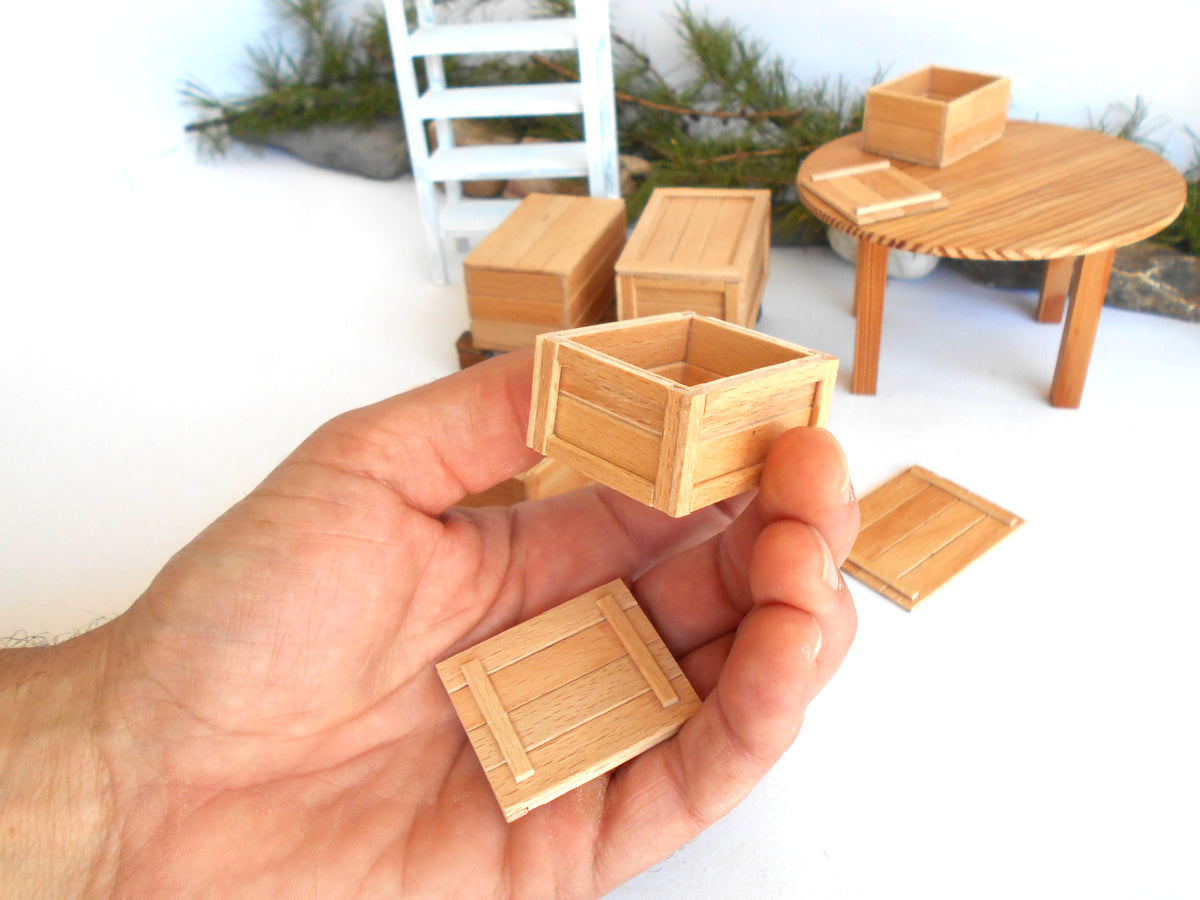 A hand holds a miniature wooden crate box with its lid placed below it, with several matching small wooden boxes and a round table in the blurred background.