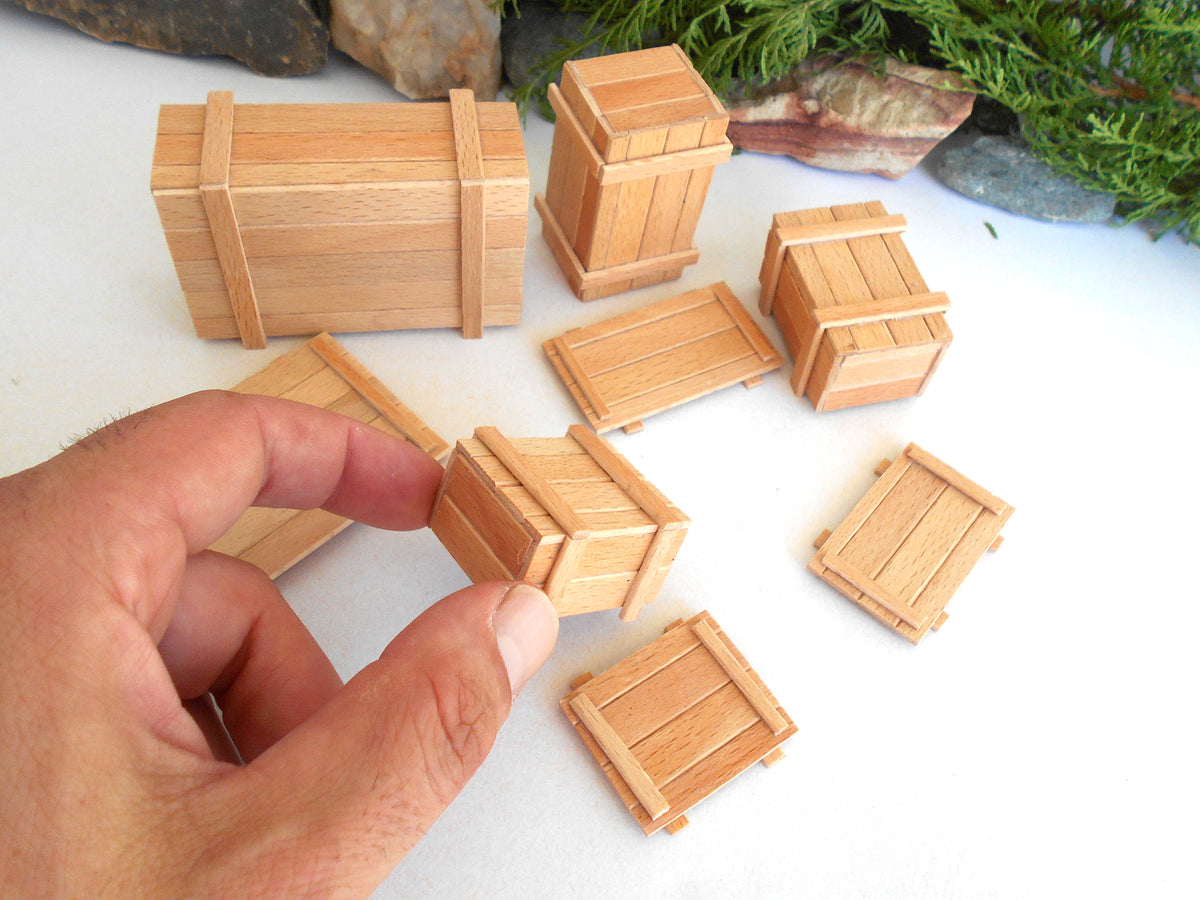 A hand holding a small wooden crate while several miniature wooden crates and boxes are arranged on a white surface with decorative rocks and greenery in the background.