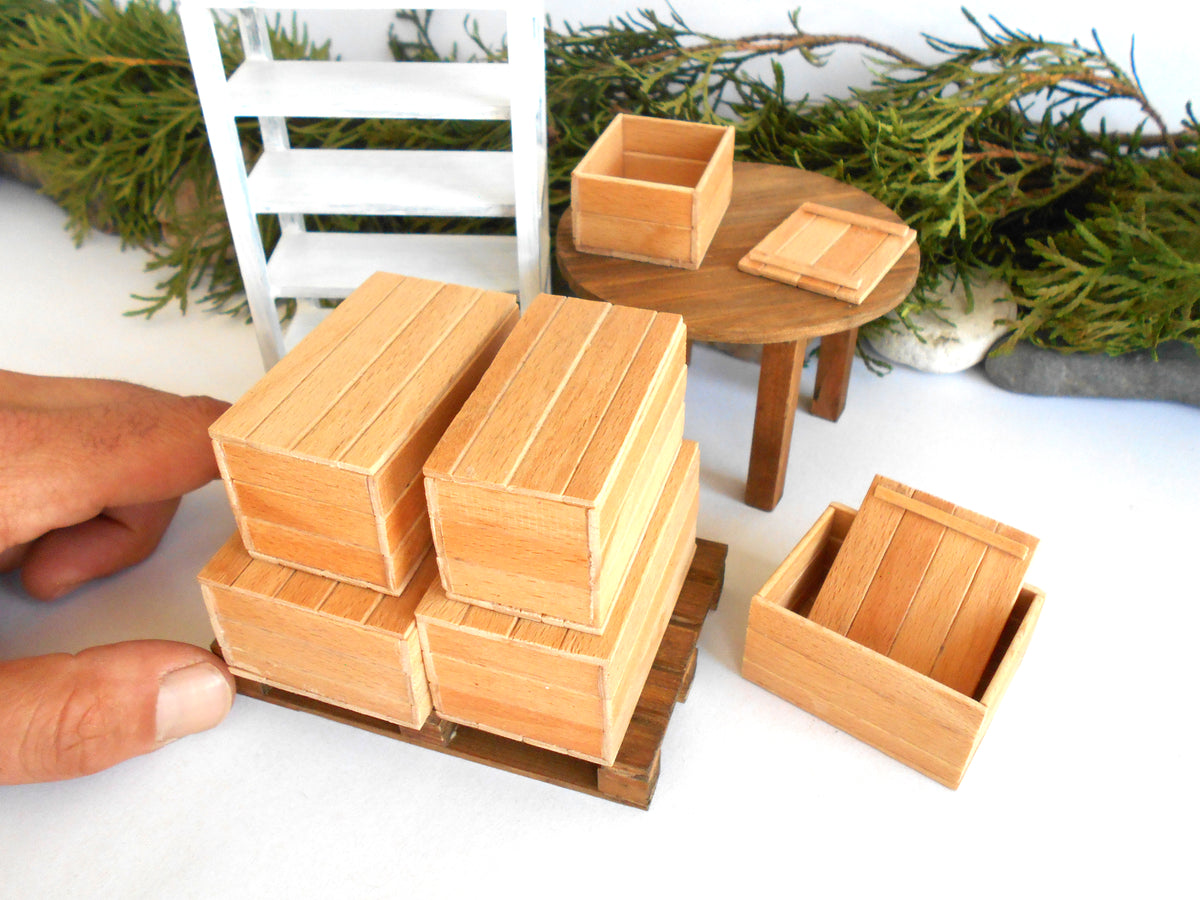 A miniature wooden crate set for dollhouse scale, featuring several light brown wooden boxes of varying sizes, with a hand holding one of the crates to show scale. The set includes a small round wooden table, a white miniature chair, and green pine branches in the background.