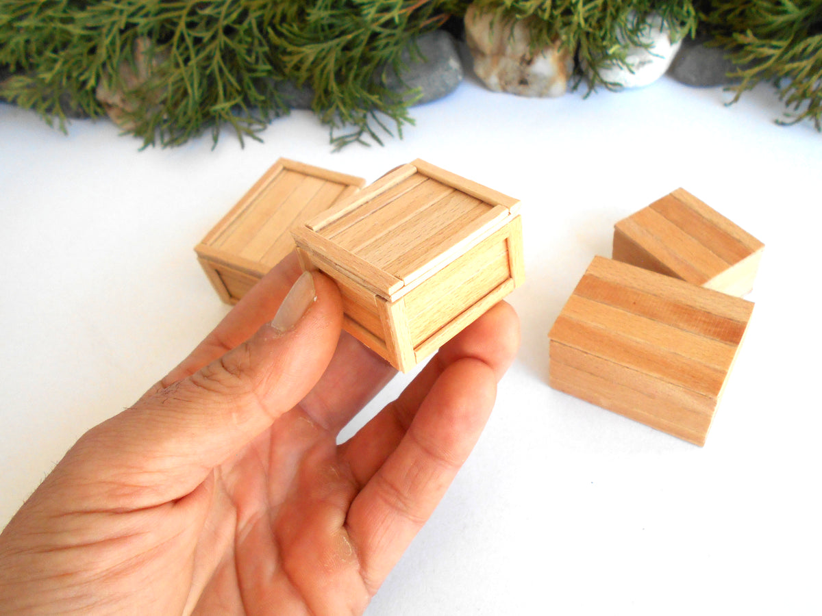 A hand holding a small wooden crate-shaped box, with several matching miniature wooden boxes scattered on a white surface in the background.