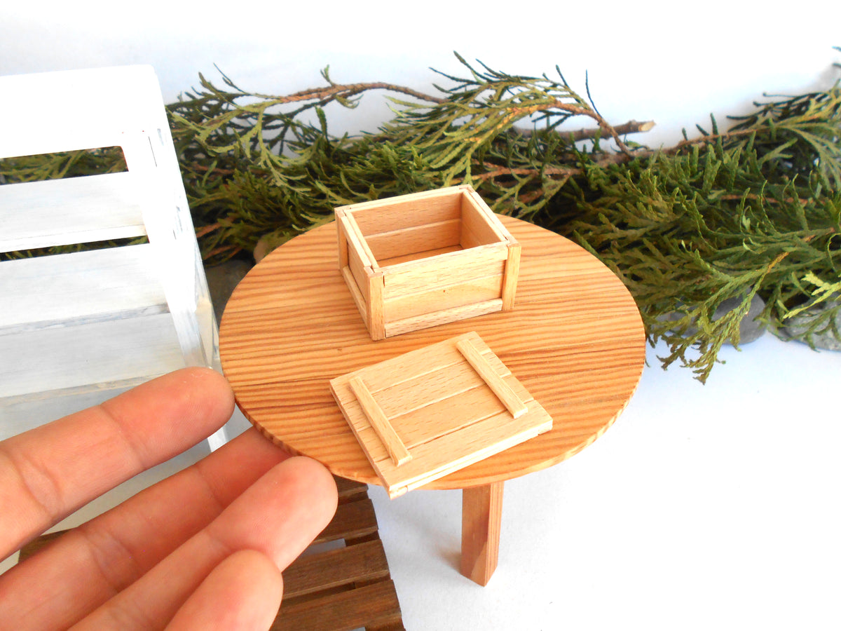 A miniature wooden crate and matching lid displayed on a small round wooden table, with a human hand visible for scale and greenery in the background.