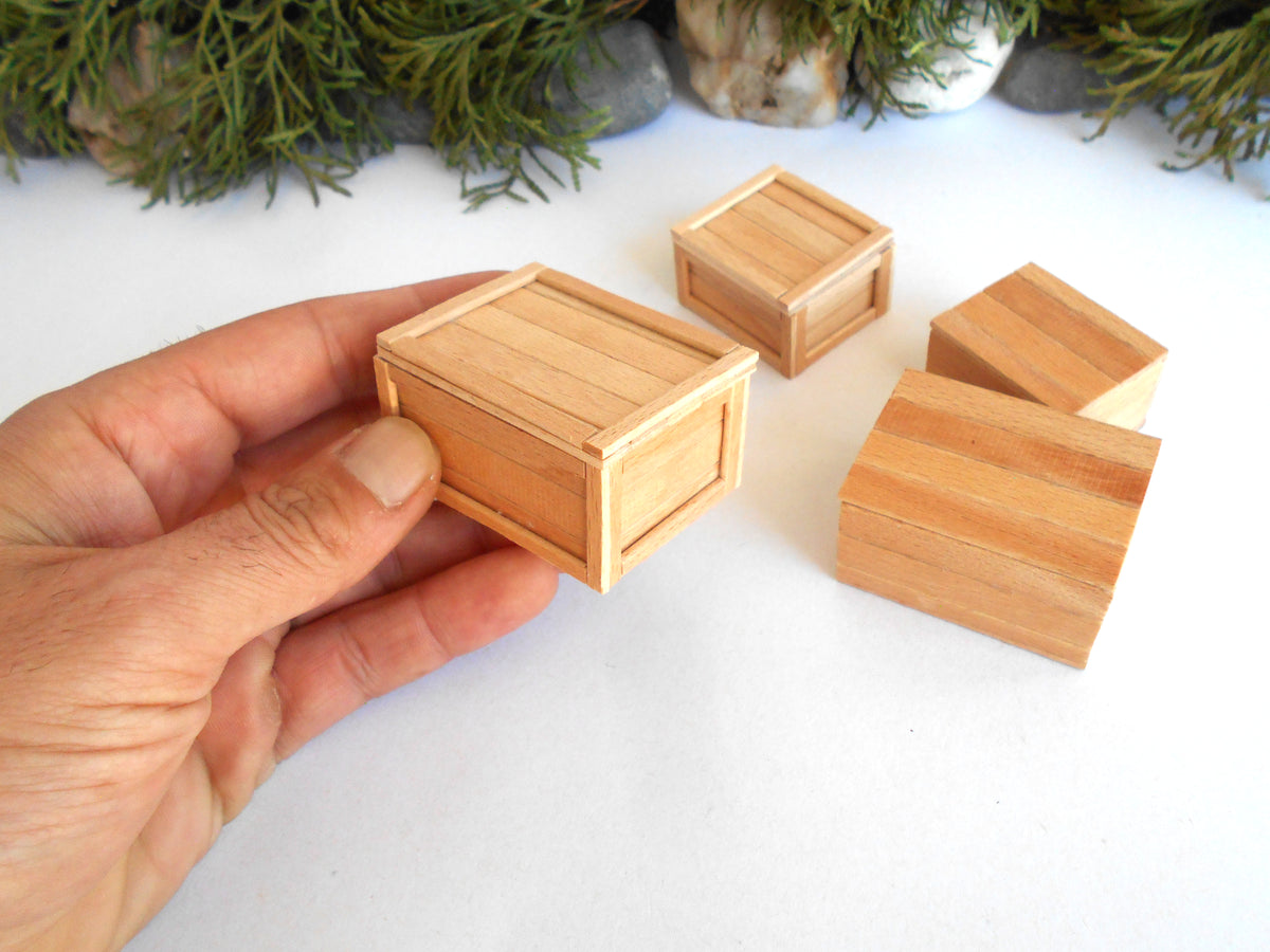 A hand holding a miniature light wood crate, with four matching miniature wooden crates arranged on a white surface in the background.