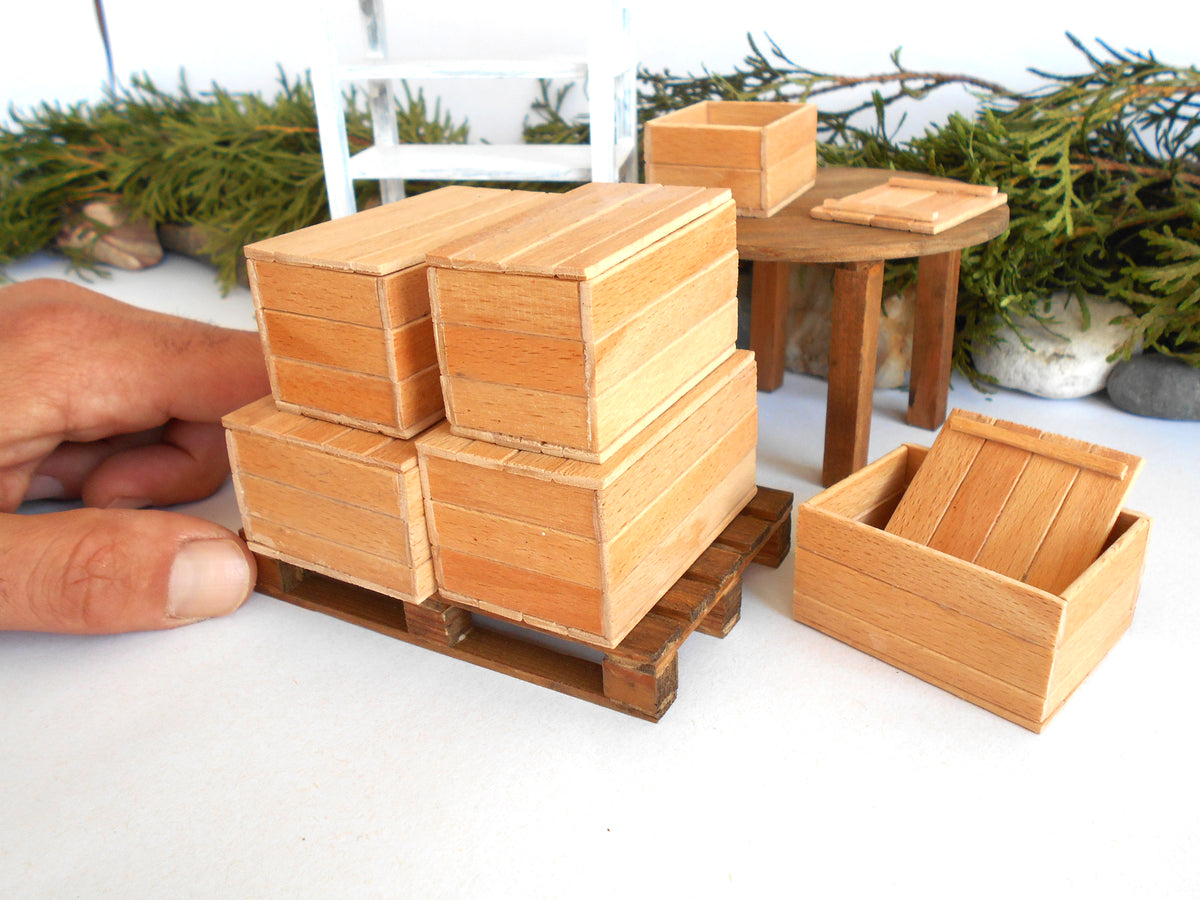 A miniature wooden box set for scale models, featuring a stack of four boxes on a small wooden pallet being held by a hand, a separate open box, and a miniature round table in the background.