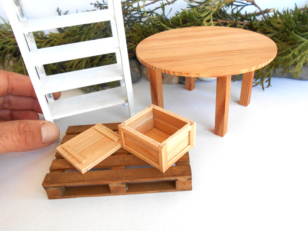 A miniature dollhouse furniture set featuring a round wooden table, a wooden crate with its lid, and a wooden pallet, shown alongside a white miniature shelf. A human hand is visible holding the pallet to demonstrate scale.