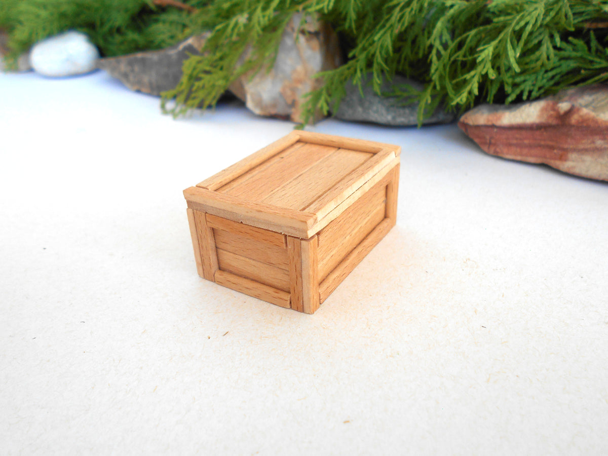 A miniature light brown wooden box with a slatted front and lid, set on a white textured surface with blurred green foliage and rocks in the background.
