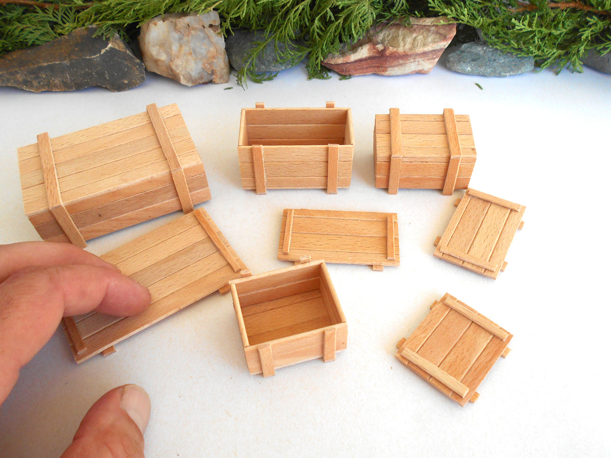 A set of miniature wooden crates and boxes in natural light wood, with a hand holding one crate lid on a white surface with decorative rocks and greenery in the background.