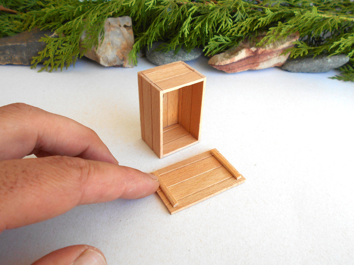 A miniature wooden box with its lid placed beside it on a white surface, with a person's fingers holding the lid. Green foliage and small rocks are visible in the background.