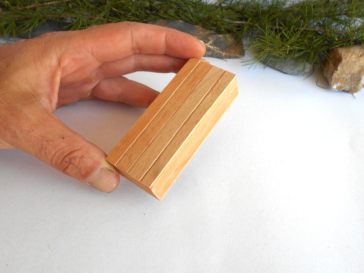 A hand holding a small rectangular wooden block with three parallel grooves, set against a white background with pine branches and small stones at the top.