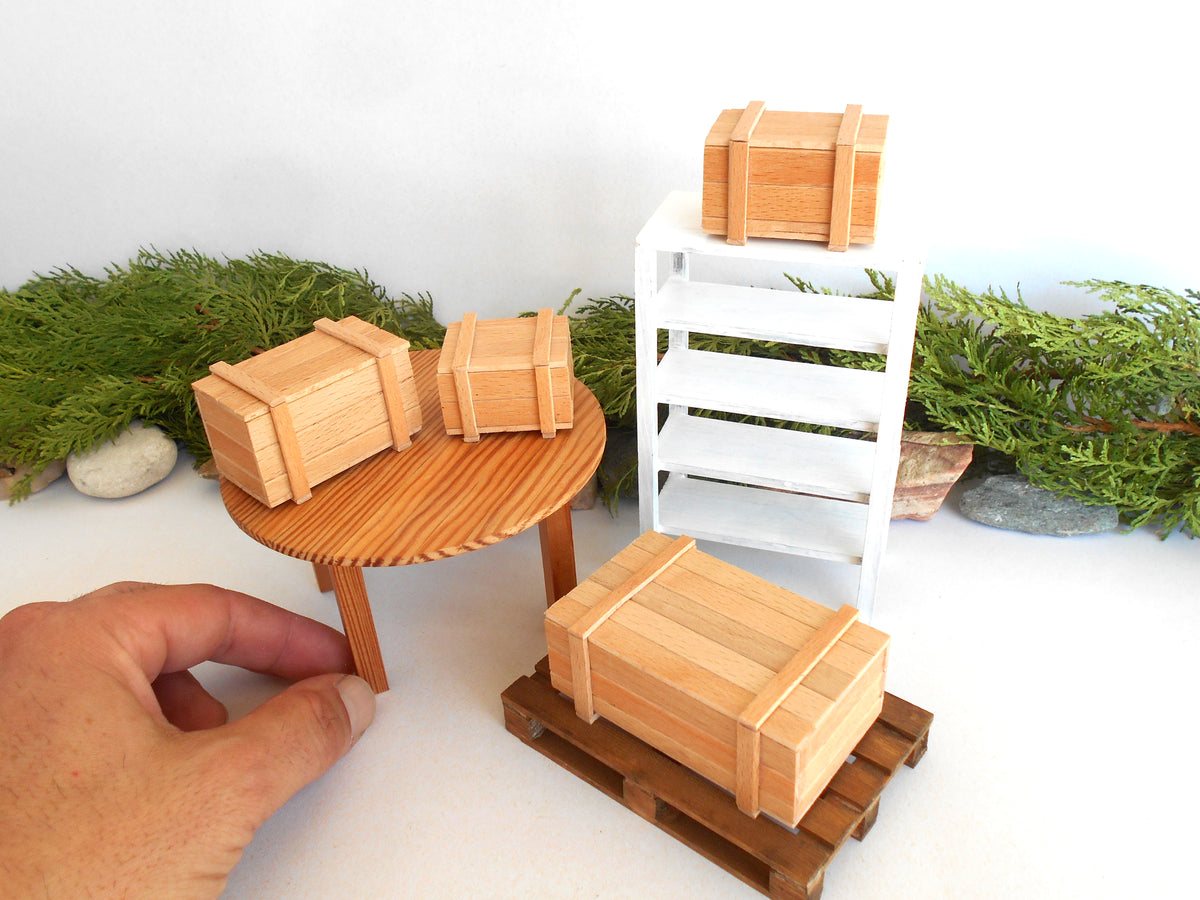 A miniature wooden dollhouse set featuring small crates, a round table, a white shelving unit, and a hand holding one of the crates to show scale, set against a white background with green foliage.