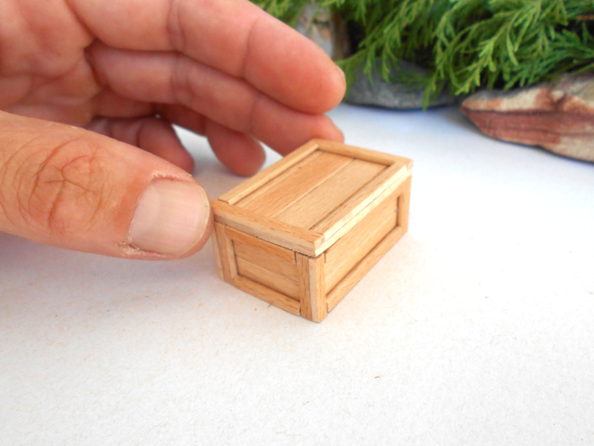A miniature light wood crate model being held by a person's hand on a white surface with a blurred green background.