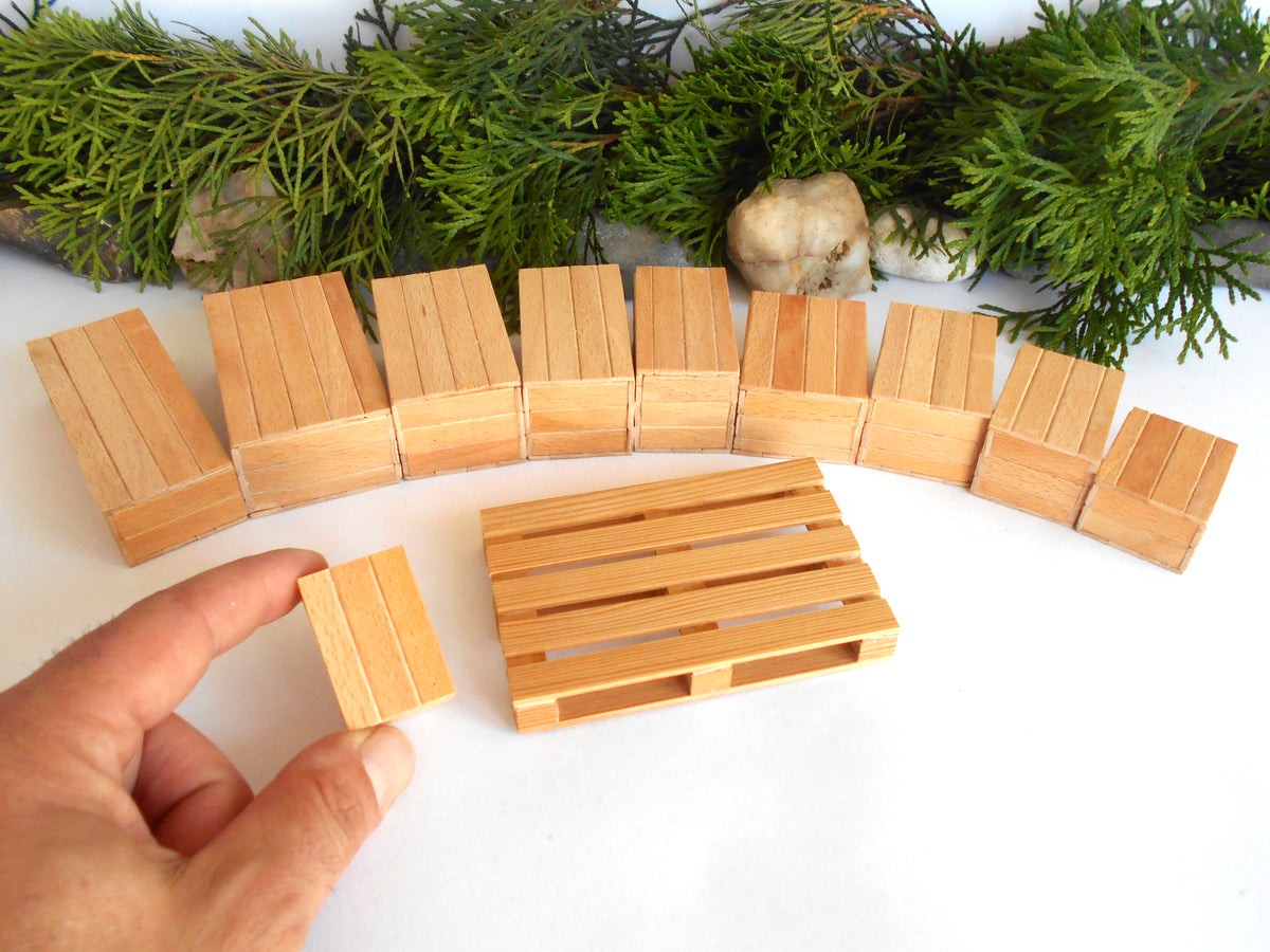 A set of miniature light brown wooden toy boxes and a small wooden pallet, with a hand holding one block in the foreground, set against a white background with green pine branches at the top.