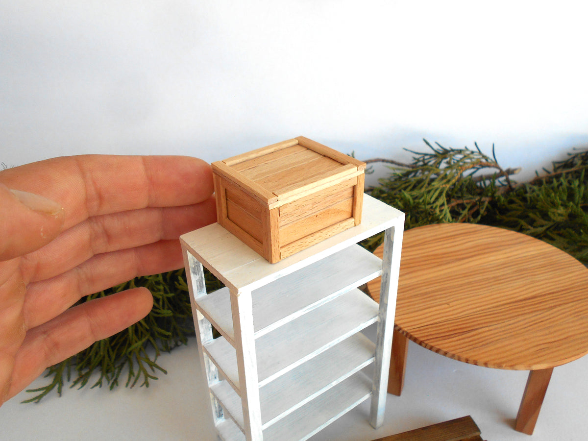 A miniature wooden storage box with a slatted lid, displayed on a small white shelving unit. A human hand is visible on the left for scale, with a miniature round table and greenery in the background.