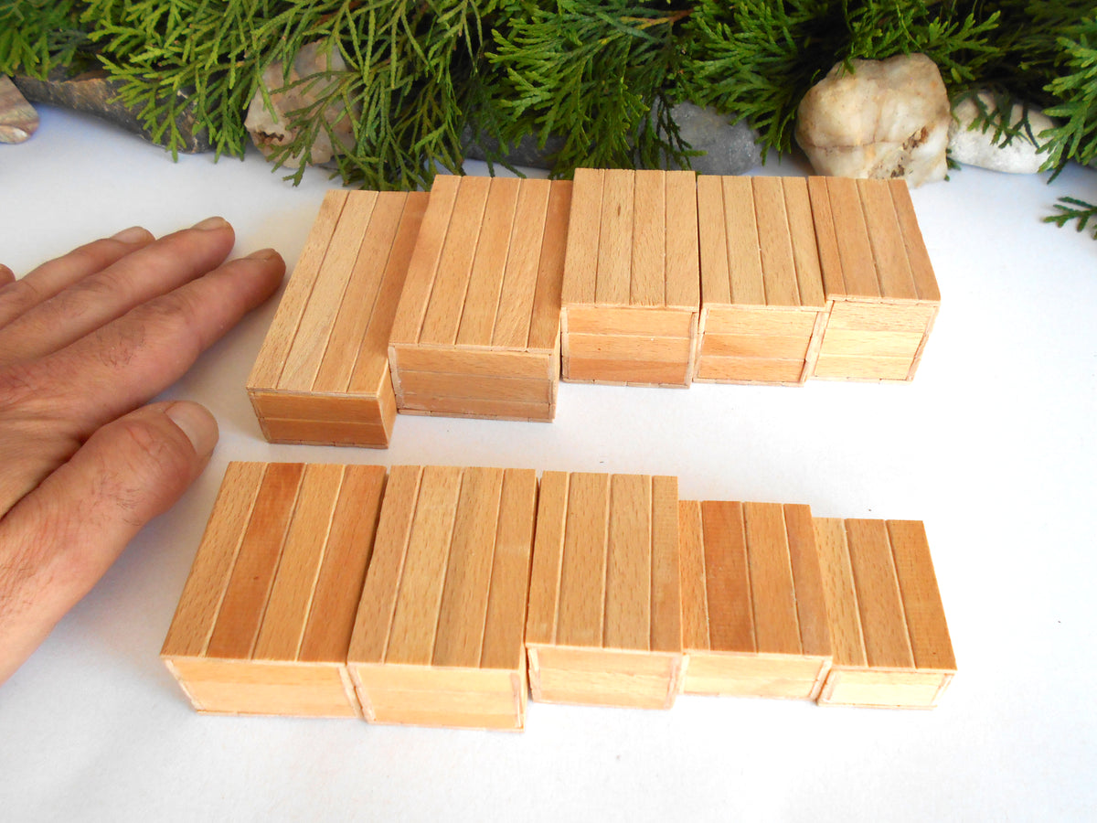 A set of light natural wood rectangular boxes arranged in two rows on a white surface, with a person's hand visible on the left for scale and green foliage in the background.