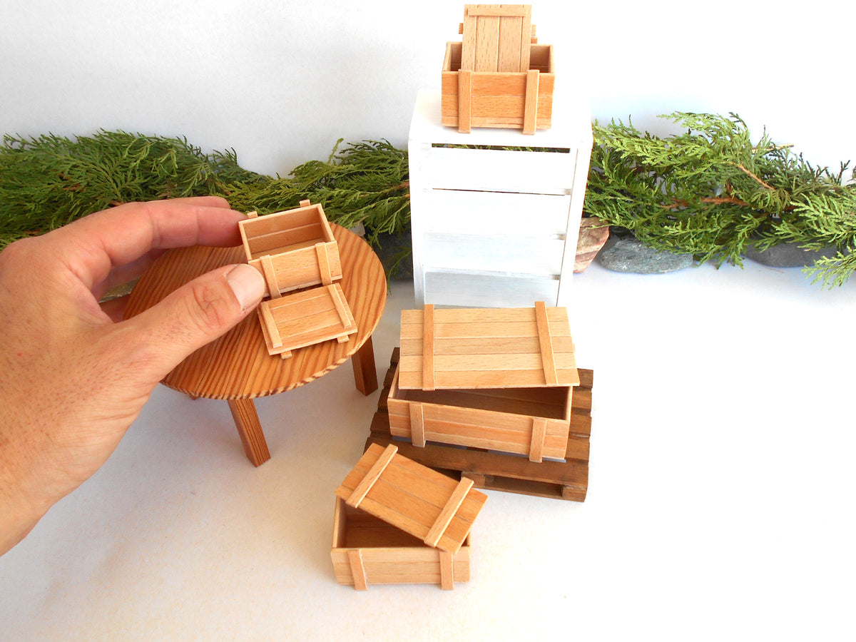 A hand holds a miniature wooden crate next to a small round table, with several other miniature wooden storage crates and a white cabinet arranged on a white surface with greenery in the background.
