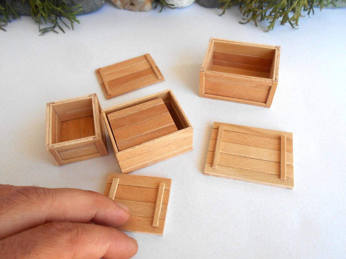 A set of miniature light wood boxes and lids, including one being held by a hand, arranged on a white surface with small green plants in the background.