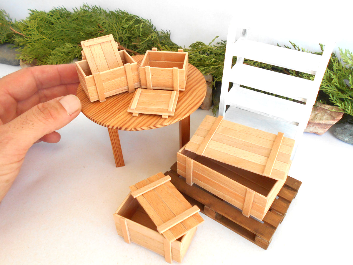 A set of miniature wooden crates and a small round wooden table, shown with a hand holding one crate to indicate scale. The scene is styled with a white slatted chair and green foliage in the background.
