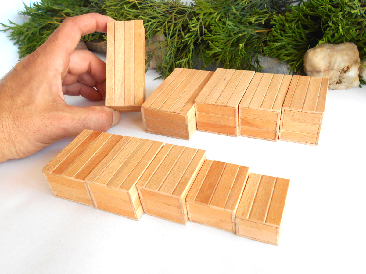A hand holds a small wooden box while several matching light wood rectangular boxes are arranged on a white surface, with greenery and a stone in the background.