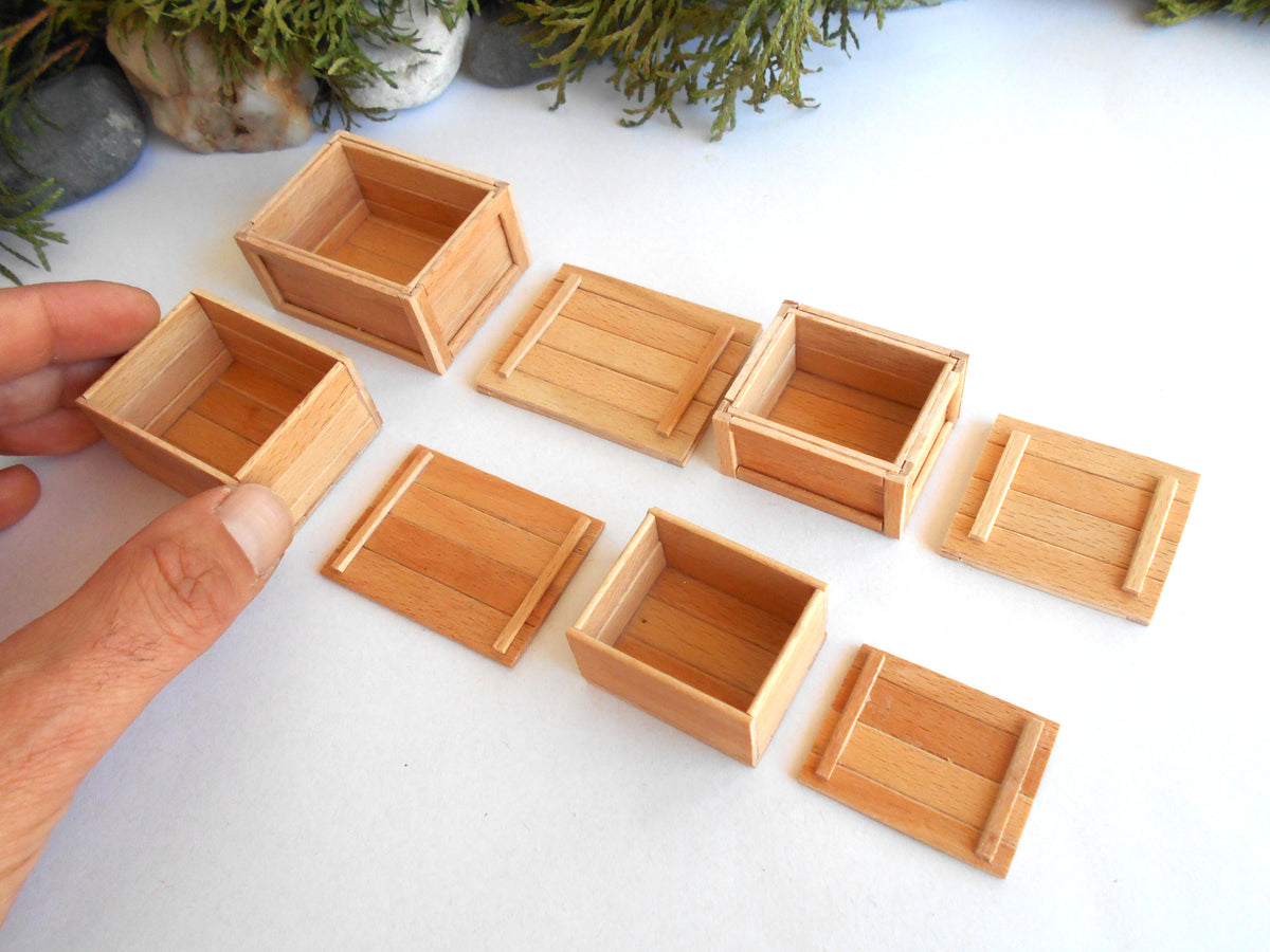 A hand holding a small wooden box next to several miniature light wood storage boxes and lids arranged on a white surface, with small potted plants in the background.