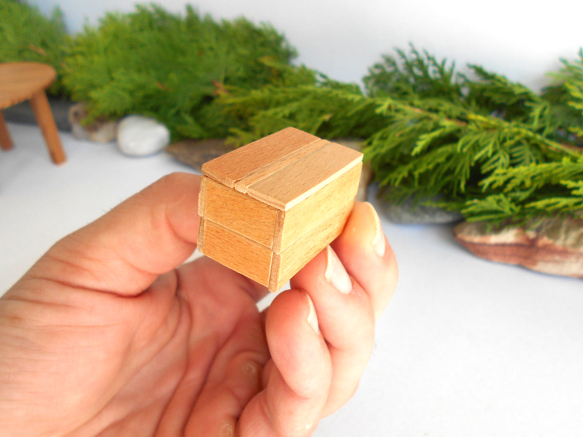 A hand holding a miniature light brown wooden box with a visible grain pattern, set against a soft-focus background of green foliage and a white surface.