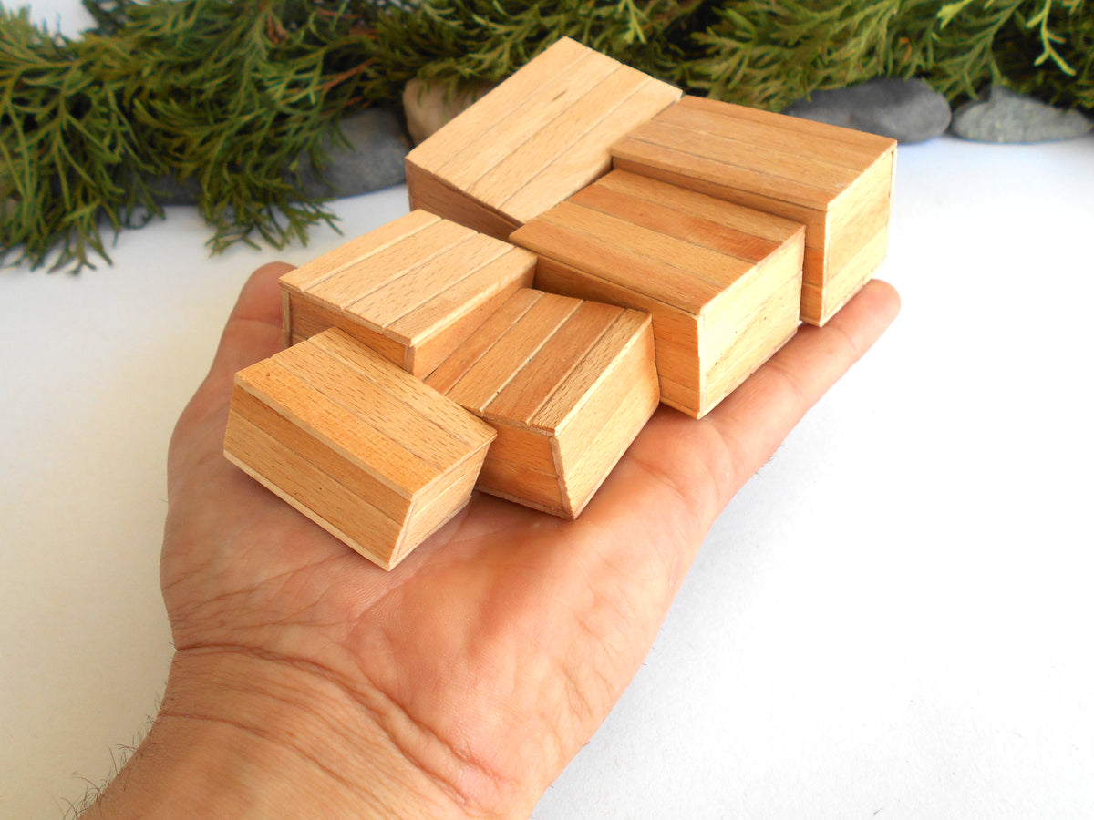A hand holding several small unfinished wooden miniature boxes with visible wood grain, set against a white background with green foliage and stones at the top.