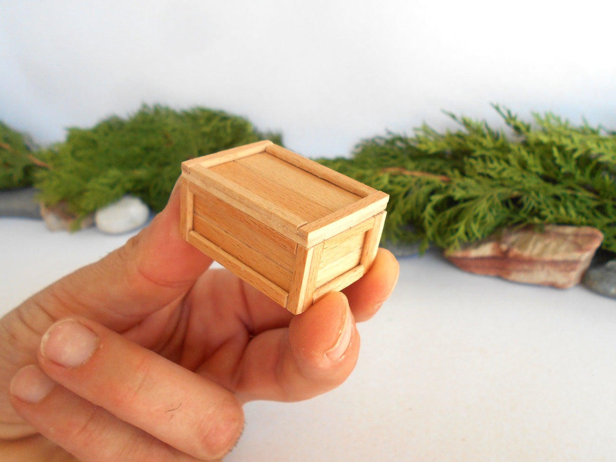 A hand holding a miniature light wood crate, shown in close-up against a soft-focus background of green foliage and white surface.