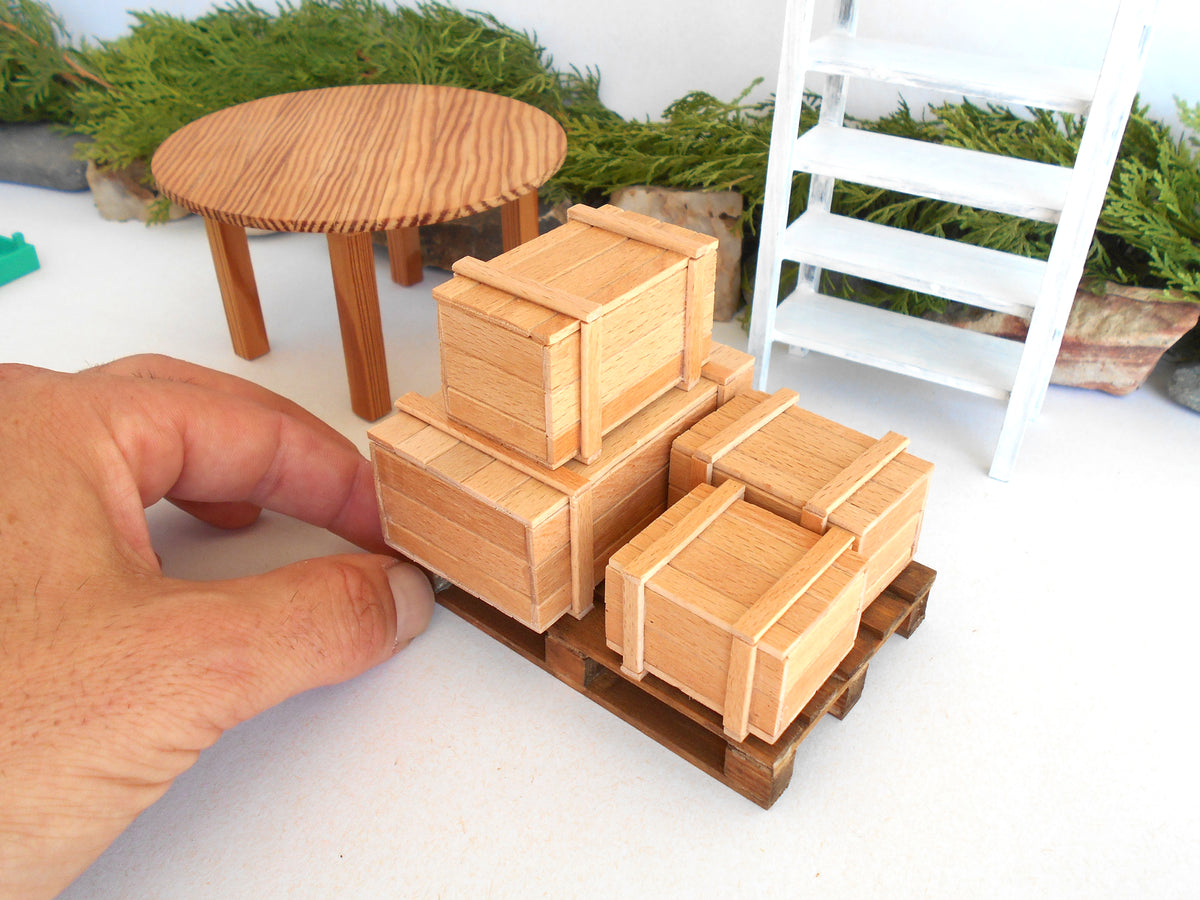 A close-up of a miniature wooden crate set on a small wooden pallet, with a hand adjusting one crate. The scene includes a small round wooden table, a white ladder, and green foliage in the background.