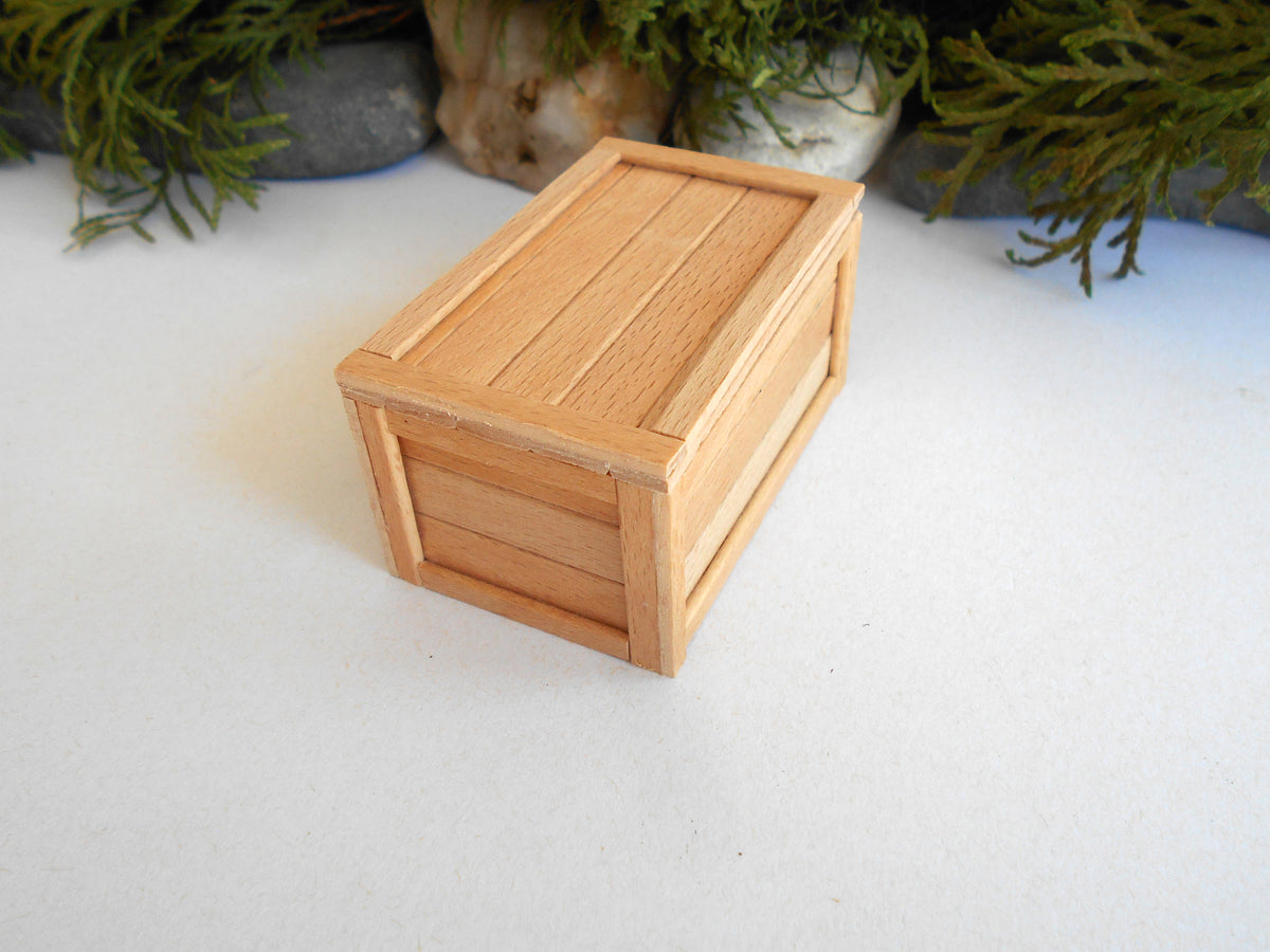 A miniature light wood box with a slatted lid and side panels, photographed on a white surface with green foliage in the background.