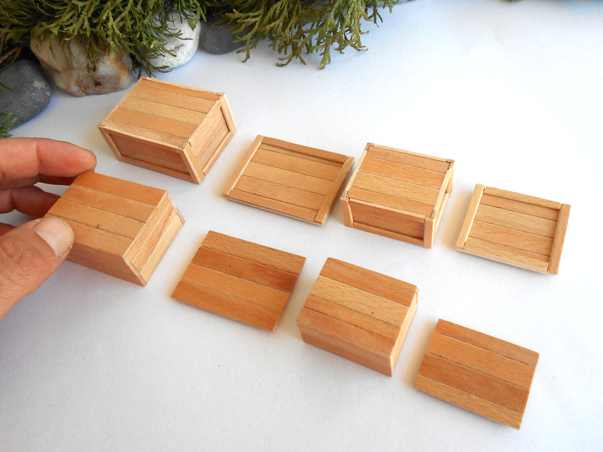 A hand holds a small rectangular wooden box on a white surface, with seven similar wooden boxes arranged around it. Moss and stones are visible in the background.