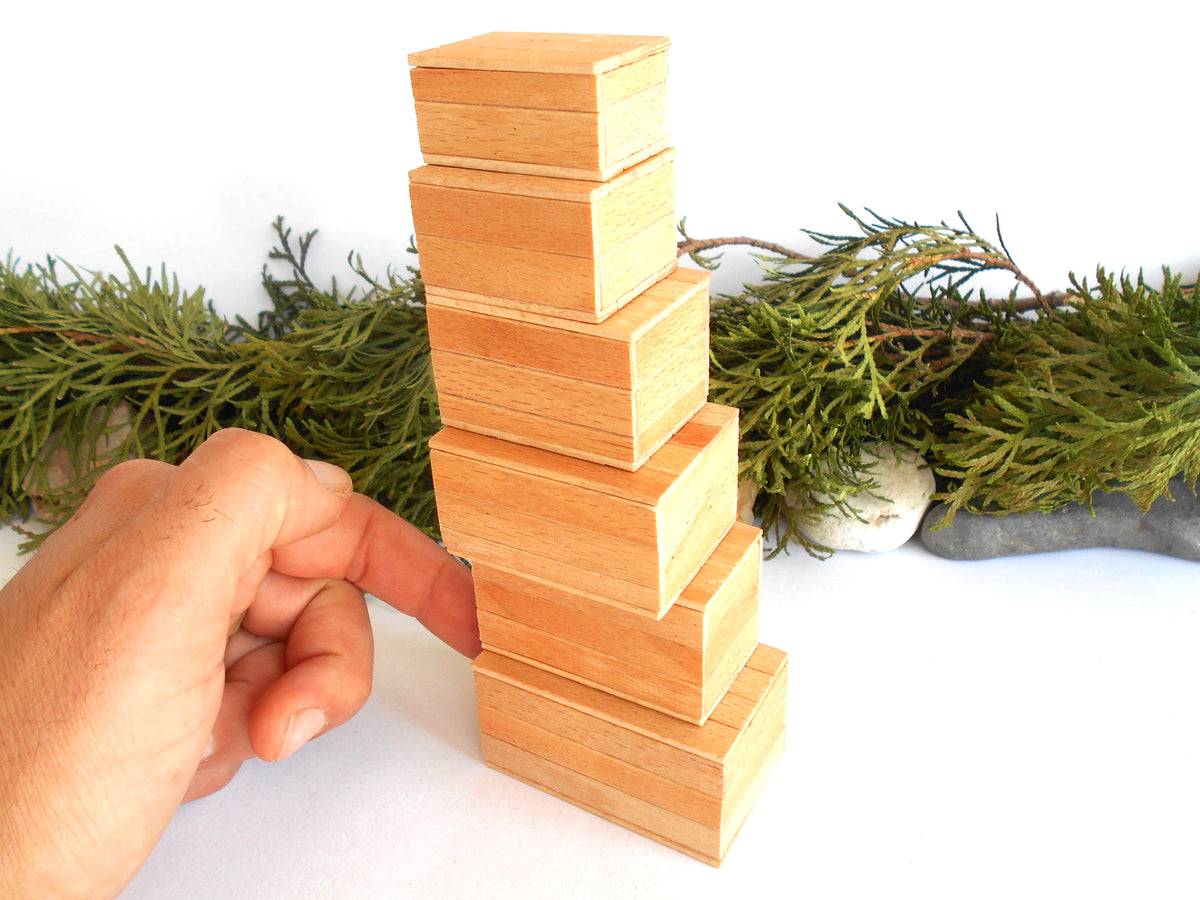 A hand is stacking light natural wood miniature boxes in a tower, with green foliage and small stones in the background against a white surface.