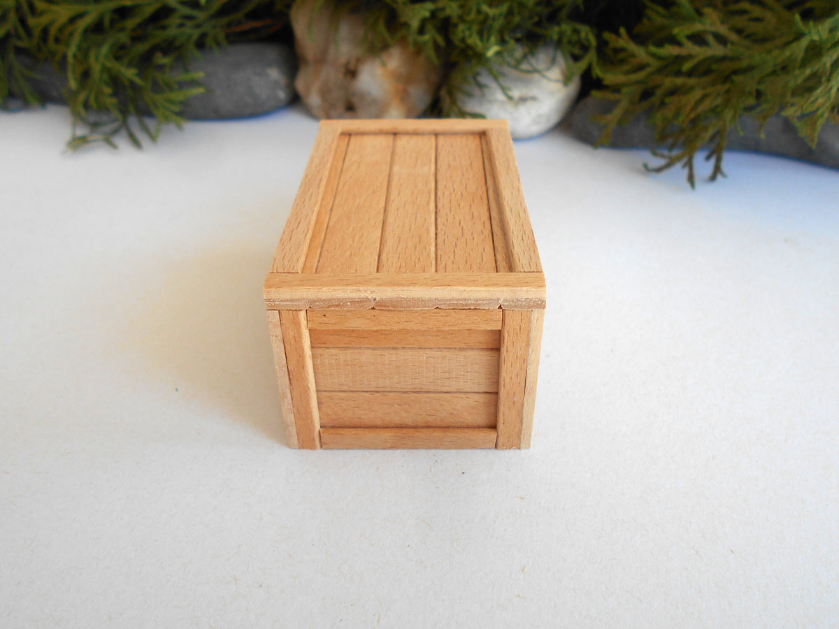 A miniature wooden storage box with a slatted lid and front panel, photographed on a white surface with blurred green foliage in the background.