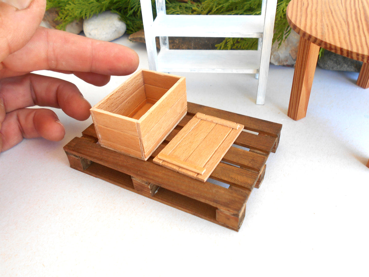A miniature wooden crate and matching lid displayed on a small wooden pallet base, with a human hand holding the crate for scale, in a tabletop miniature setup.