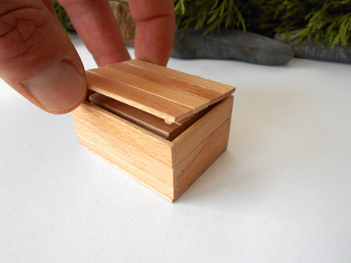 A miniature light brown wooden box with a lid, being opened by a person's fingers on a white surface with a blurred background.