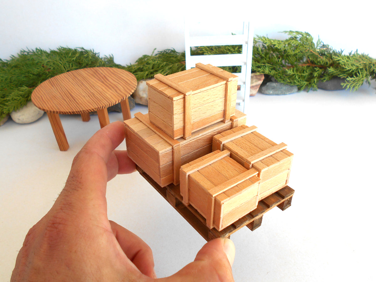 A hand holds a miniature wooden cargo pallet with several small wooden crates, with a miniature round table and white ladder in the background.