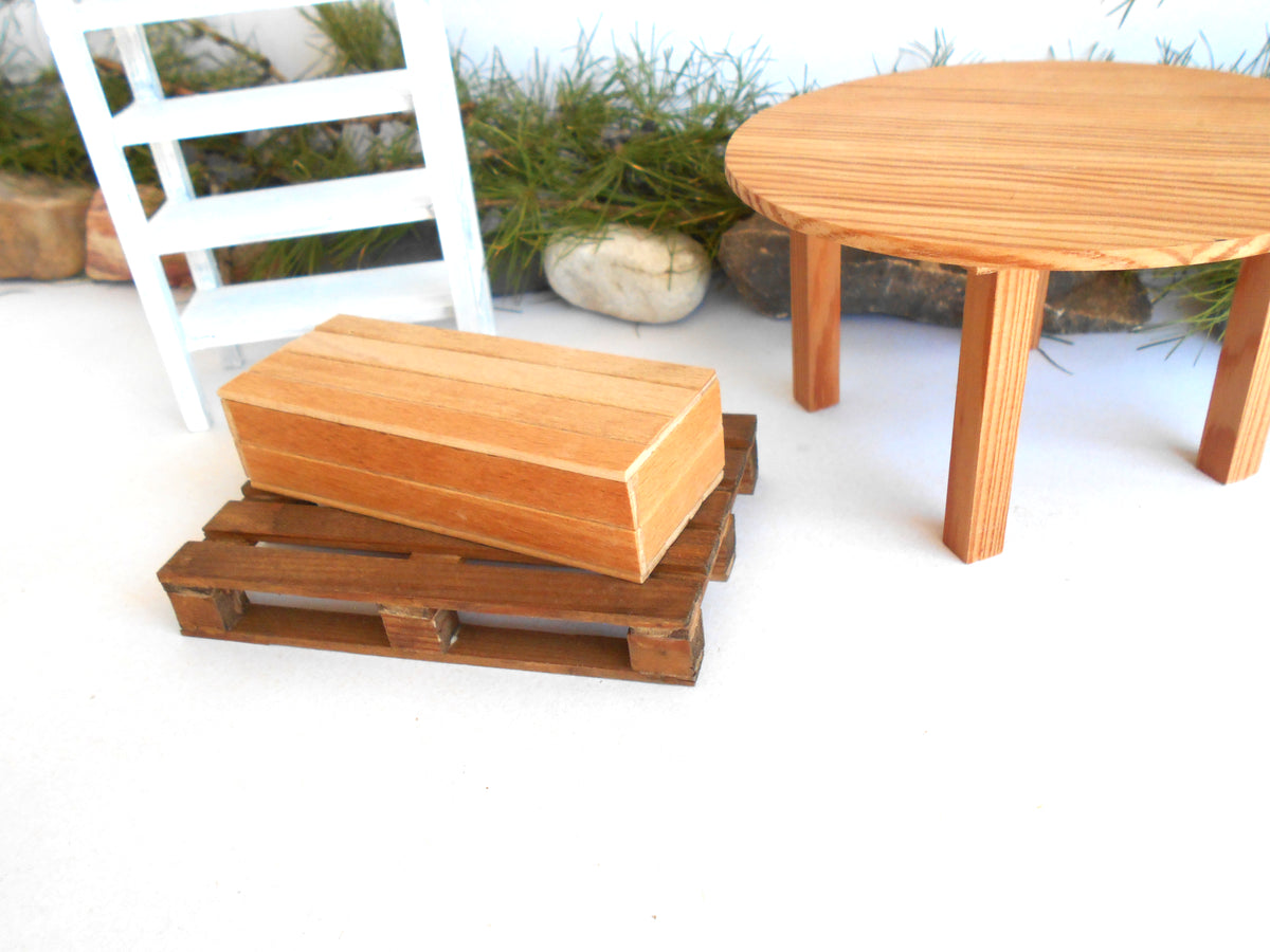 A miniature wooden dollhouse furniture set featuring a small rectangular wooden box resting on a dark brown wooden pallet, with a round light wood table in the background.