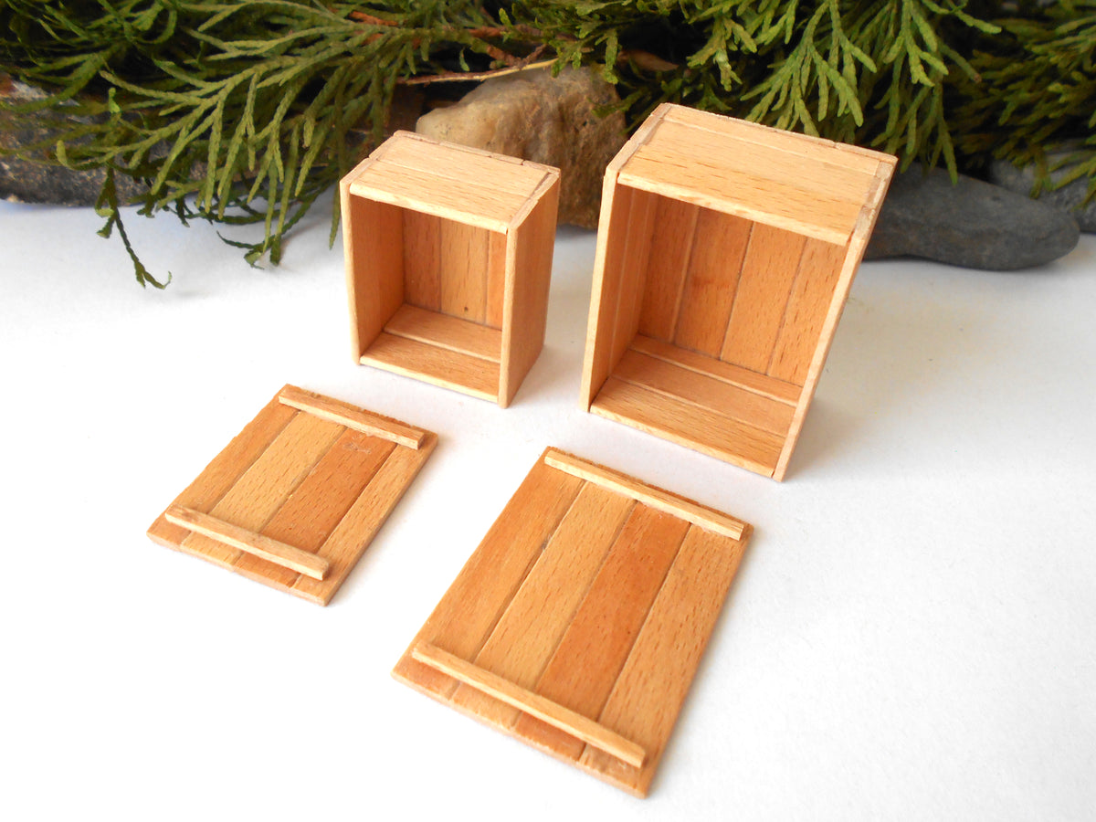 Four miniature light wood boxes and lids arranged on a white surface with green pine branches and small stones in the background.