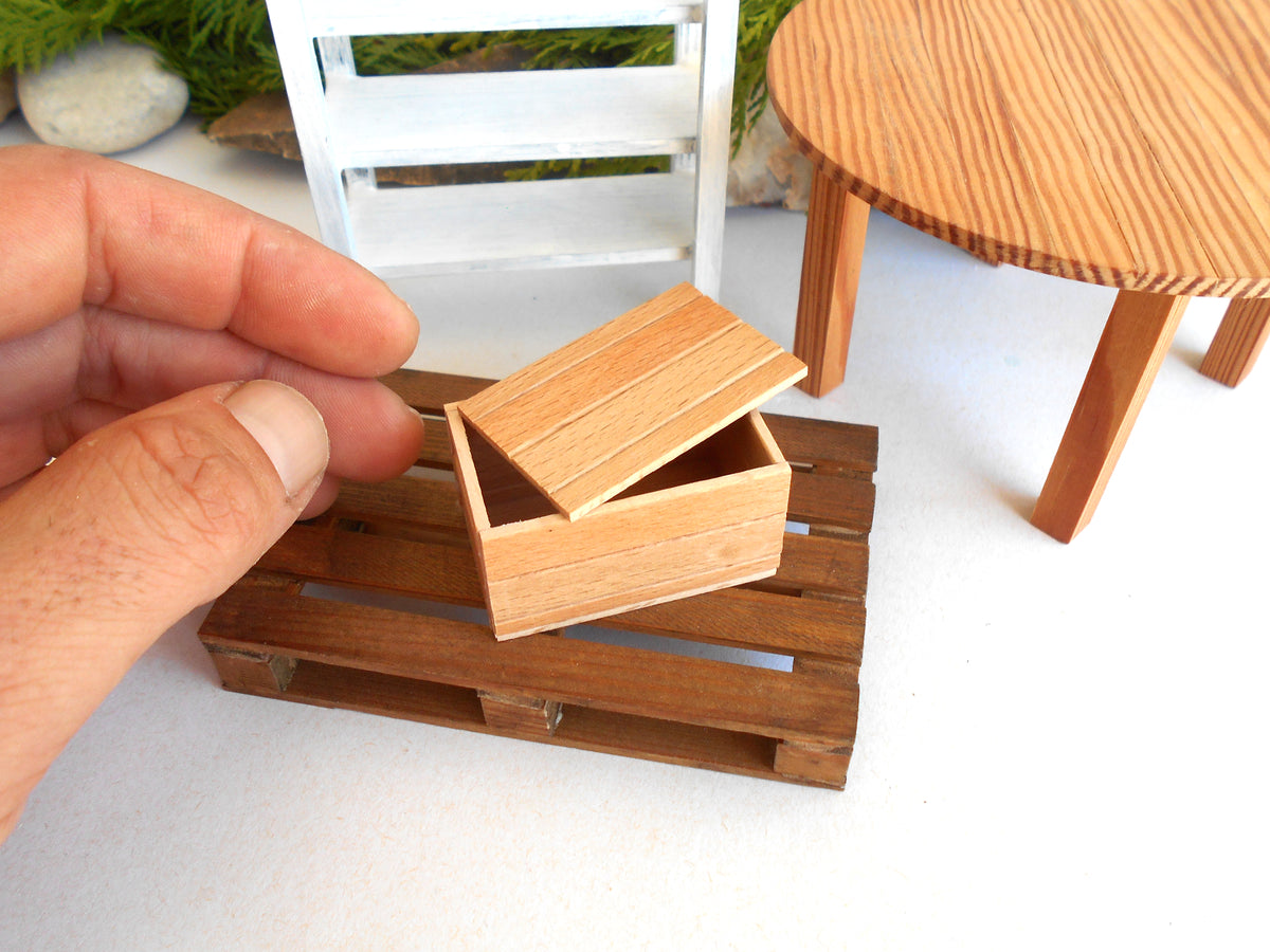 A miniature wooden storage box with its lid slightly open, resting on a small wooden pallet-like base. A human hand is holding the box to show scale, with miniature furniture and greenery in the background.